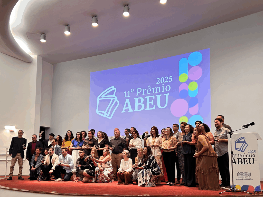A imagem mostra um grande grupo de pessoas reunidas no palco de um auditório, posando para foto coletiva. Todas seguram troféus do Prêmio ABEU. O telão ao fundo exibe a identidade visual do evento com o texto “11º Prêmio ABEU – 2025” em azul e elementos gráficos circulares coloridos. À direita, está o púlpito com o logotipo da ABEU. O ambiente é amplo, com iluminação embutida no teto e paredes claras.